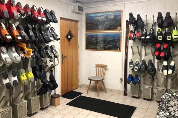 Miestnosť s lyžiarskymi topánkami na sušiakoch a obrazmi hôr na stene.