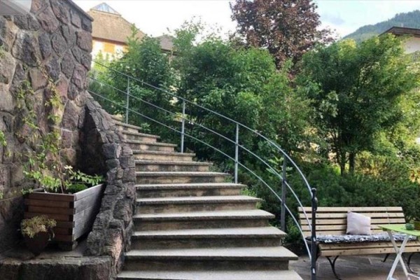 Kľukaté kamenné schody vedúce nahor medzi zelenými kríkmi, vedľa lavička a stolík.