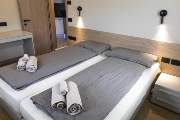 Dve jednolôžka so sivým povlečením a uterákmi, nočné stolíky a lampy na stene.