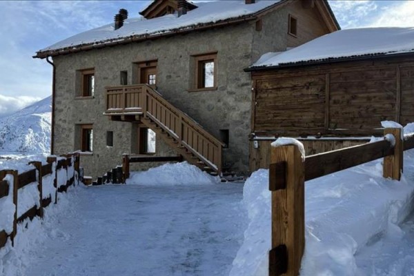Kamenný dom s zasneženou strechou a dreveným zábradlím v horskej oblasti.